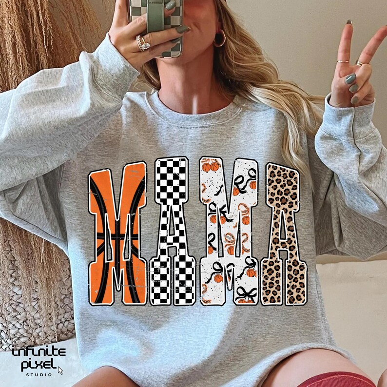 Puede incluir: Una sudadera gris claro con la palabra "MAMA" en letras grandes y decorativas. La "M" es naranja con un dise&ntilde;o de baloncesto, la primera "A" es a cuadros blancos y negros, la segunda "M" tiene un dise&ntilde;o de calabaza y lazo, y la &uacute;ltima "A" es de estampado de leopardo.