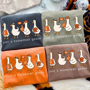 May include: Four sweatshirts in charcoal, olive green, orange, and brown. Each features a cartoon goose wearing a basketball jersey, headband, hat, and holding a foam finger or basketball. The text "just a basketball goose" is printed below.