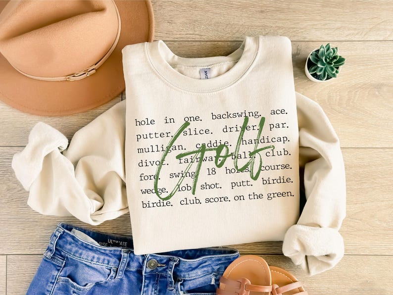 May include: A cream-colored sweatshirt with green text that reads "Golf" and a list of golf terms. The text is written in a typewriter font.