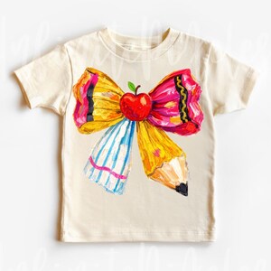 May include: Cream-colored t-shirt featuring a colorful bow design with an apple and pencil elements. The bow has yellow, pink, and black accents, with a blue and white striped section. A red apple sits in the center of the bow.