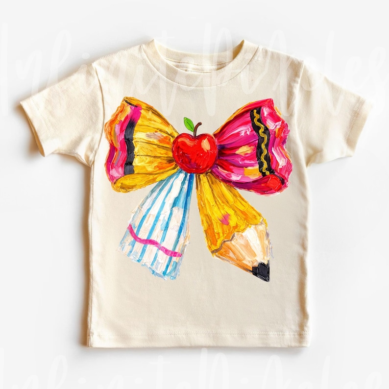 May include: Cream-colored t-shirt featuring a colorful bow design with an apple and pencil elements. The bow has yellow, pink, and black accents, with a blue and white striped section. A red apple sits in the center of the bow.