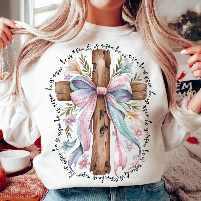 May include: White sweatshirt with a watercolor illustration of a wooden cross with a pink and blue bow and floral accents. The text "He is risen. He is risen. He is risen. He is risen. He is risen. He is risen. He is risen. He is risen." is printed around the cross.