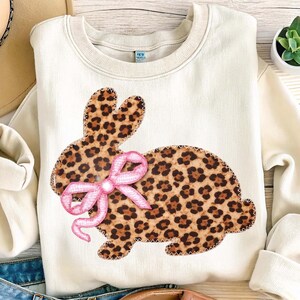 May include: Cream-coloured sweatshirt featuring a leopard print bunny design with a pink bow. The bunny is outlined with a white zig-zag stitch. The sweatshirt is a casual, comfortable style.