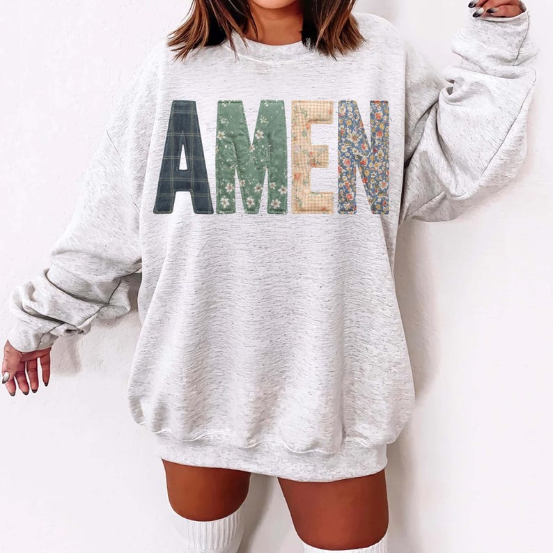 May include: A light gray sweatshirt with the word "AMEN" across the front. The letters are in a patchwork style, with plaid, floral, and gingham patterns. The sweatshirt has long sleeves and a crew neck.