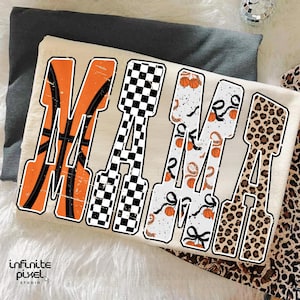 Puede incluir: Una camiseta color crema con la palabra "MAMA" en letras grandes y en negrita. Cada letra tiene un dise&ntilde;o &uacute;nico: baloncesto, a cuadros, estampado de baloncesto y serpiente, y estampado de leopardo. La camiseta est&aacute; doblada sobre una superficie gris.