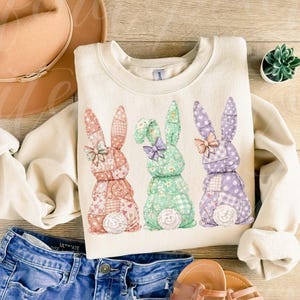 May include: Cream-colored sweatshirt featuring three patchwork bunny rabbits in pastel colors. The bunnies have patterned ears and bows. The sweatshirt is paired with blue jeans, a hat, and sandals, suggesting a spring or Easter theme.