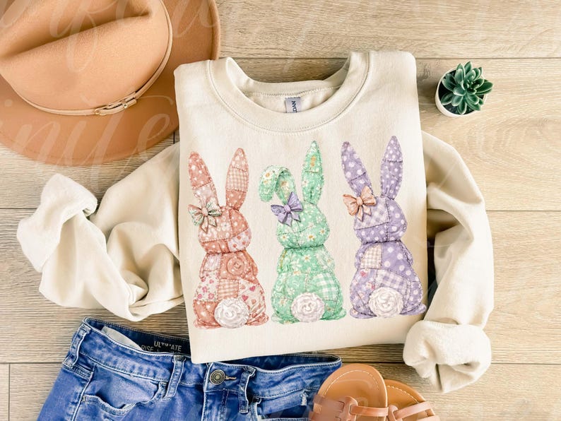 May include: Cream-colored sweatshirt featuring three patchwork bunny rabbits in pastel colors. The bunnies have patterned ears and bows. The sweatshirt is paired with blue jeans, a hat, and sandals, suggesting a spring or Easter theme.