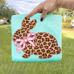 May include: A turquoise plush square featuring a leopard print bunny applique. The bunny has a pink bow and is stitched with brown thread. The image is held by a hand with a gold bracelet and purple nail polish.