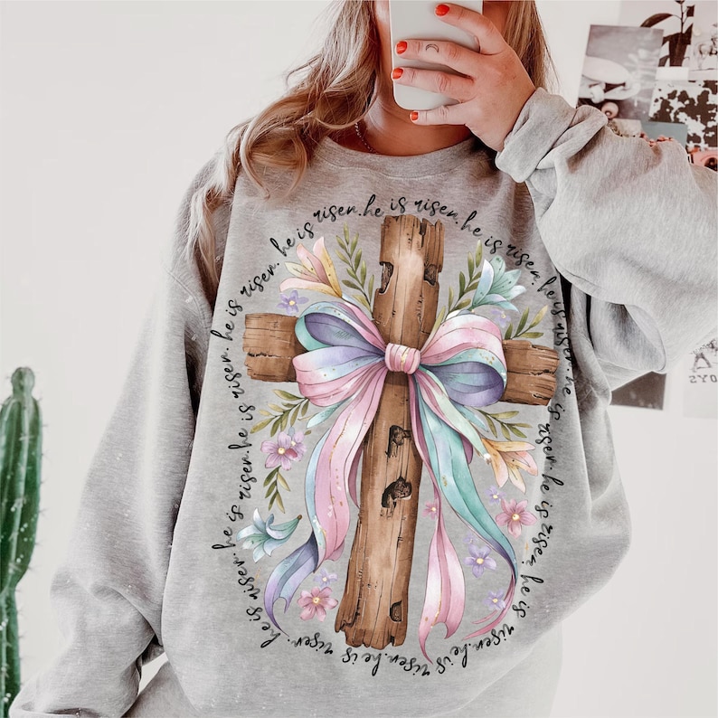 May include: Gray sweatshirt with a wood cross and a pink and blue bow. The cross is surrounded by flowers and the text "He is risen. He is risen. He is risen. He is risen. He is risen. He is risen. He is risen. He is risen." is printed around the cross.