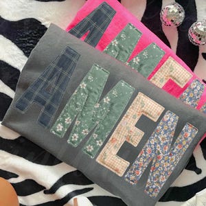 May include: Two decorative pillows, one gray and one pink, with the word "AMEN" appliqued on the front. The letters are made of various patterned fabrics, including plaid, floral, and gingham. The pillows rest on a zebra-print surface.