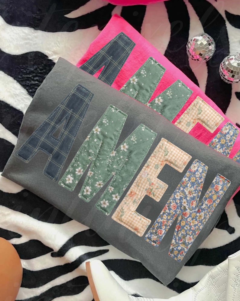 May include: Two decorative pillows, one gray and one pink, with the word "AMEN" appliqued on the front. The letters are made of various patterned fabrics, including plaid, floral, and gingham. The pillows rest on a zebra-print surface.