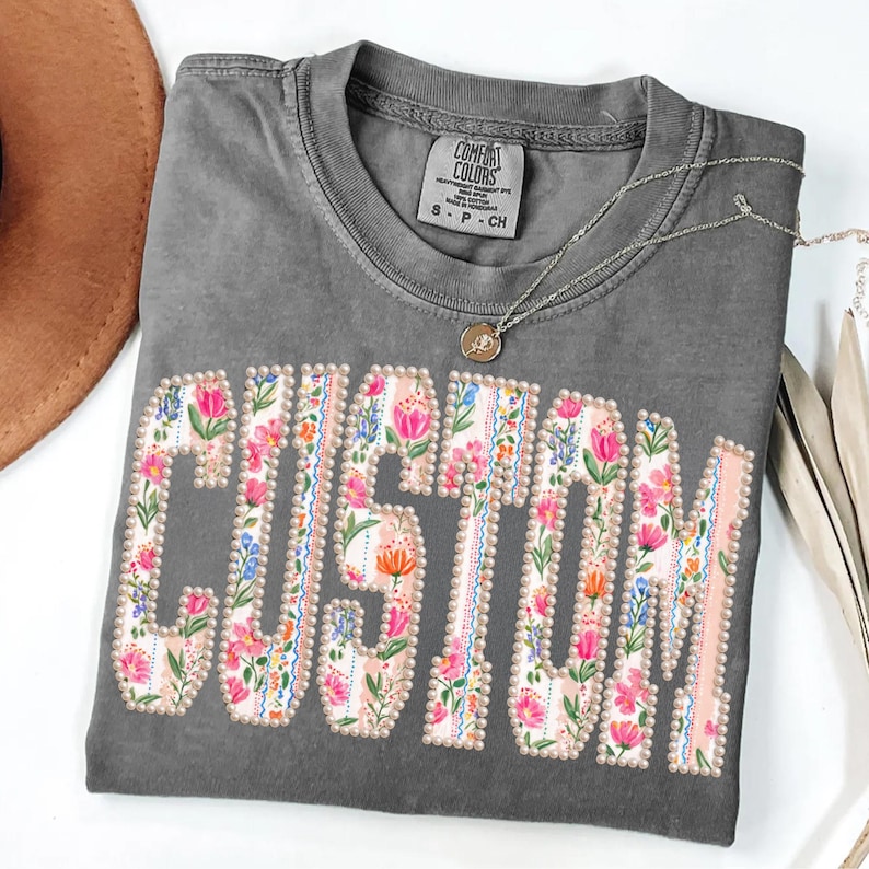 Puede incluir: Camiseta gris Comfort Colors con la palabra "CUSTOM" en un dise&ntilde;o floral. Las letras est&aacute;n delineadas con peque&ntilde;as perlas y rellenas de flores rosas, azules y naranjas. Tambi&eacute;n se ve un collar plateado con un colgante.