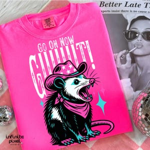 May include: A bright pink t-shirt with the text "GO ON NOW GIT!" and a graphic of a possum wearing a cowboy hat and bandana. The possum is black, white, and teal. The shirt has a Comfort Colors tag.