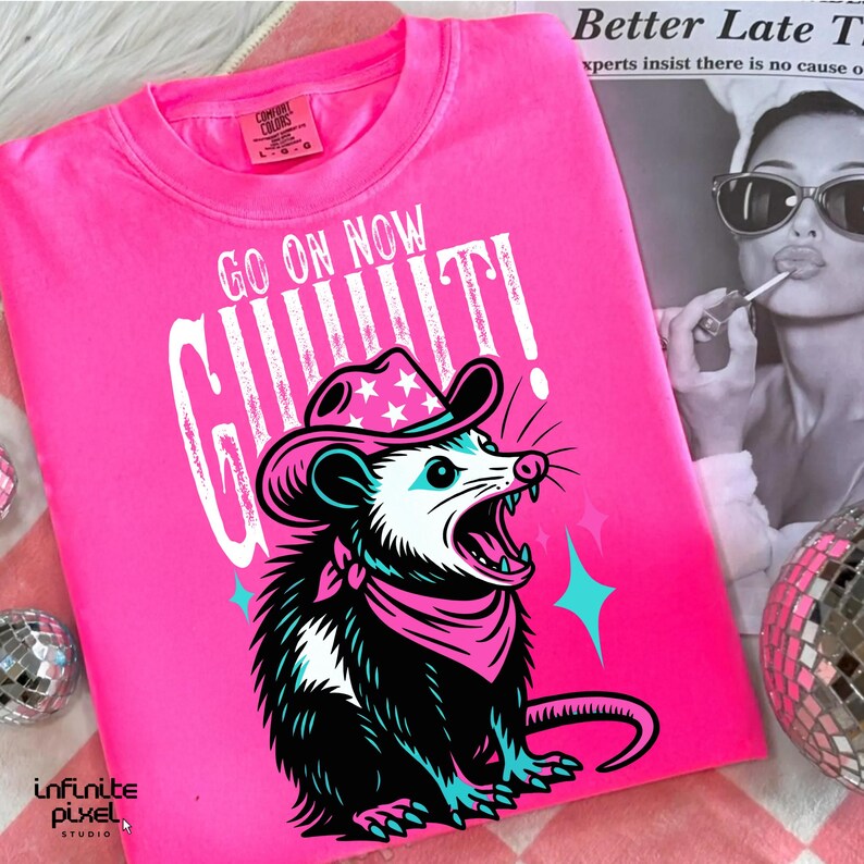 May include: A bright pink t-shirt with the text "GO ON NOW GIT!" and a graphic of a possum wearing a cowboy hat and bandana. The possum is black, white, and teal. The shirt has a Comfort Colors tag.