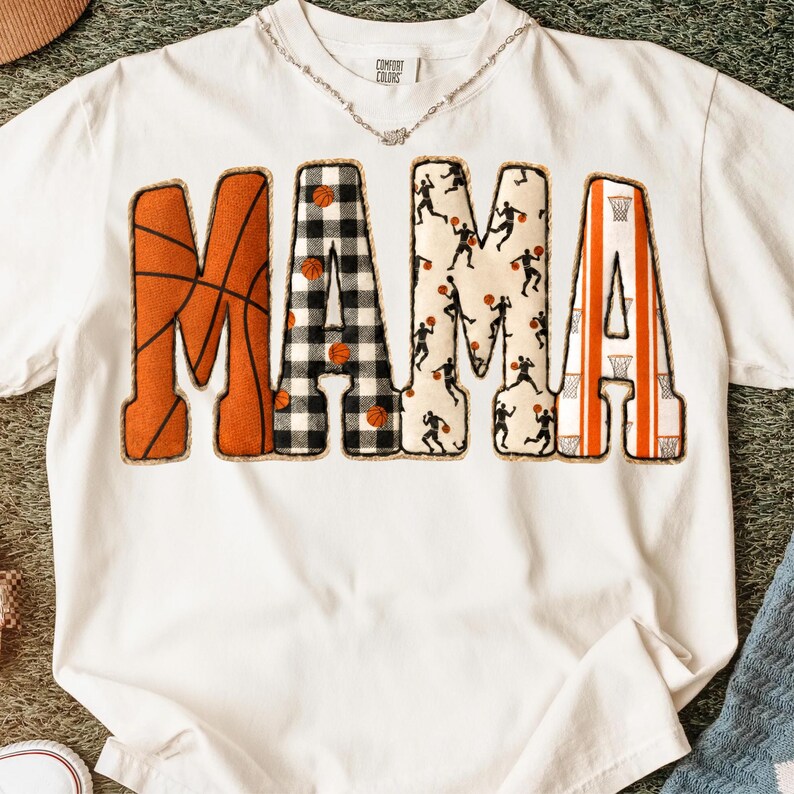 Puede incluir: Camiseta blanca con la palabra "MAMA" en letras decorativas. La "M" tiene un dise&ntilde;o de baloncesto, las "A"s con aros de baloncesto y la "A" central con un dise&ntilde;o a cuadros blanco y negro.