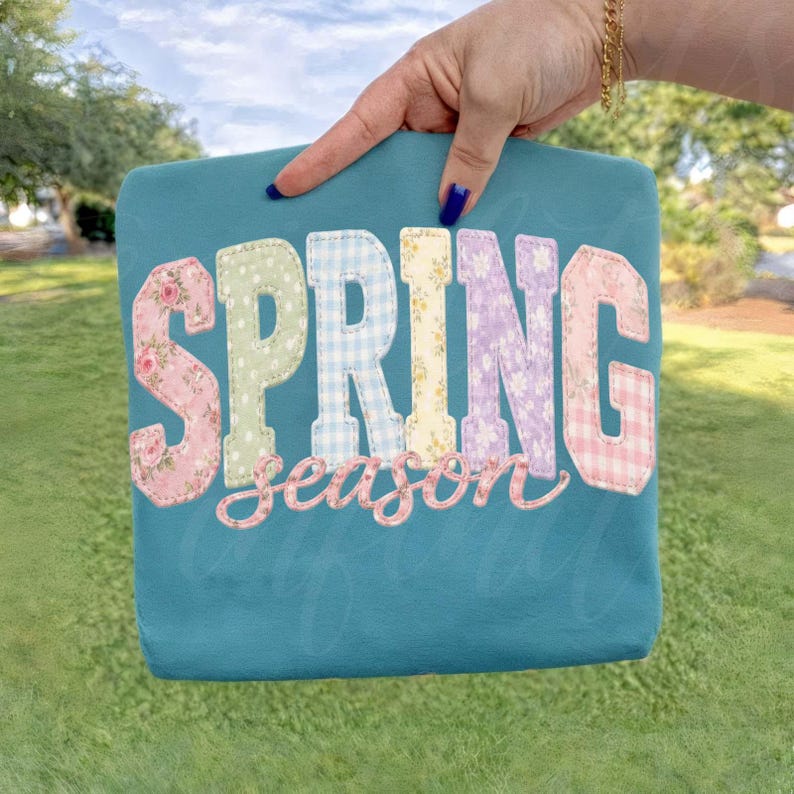 May include: Teal-colored tote bag with the words "SPRING season" appliqued in colorful floral, gingham, and patterned fabrics. The bag is held up by a hand with blue nail polish, against a blurred green background.
