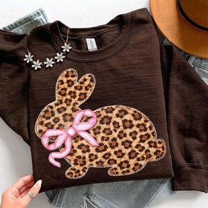 May include: A brown sweatshirt with a leopard print bunny design and a pink bow. The bunny is outlined with white stitching. A silver necklace with flower-shaped pendants is draped on the sweatshirt.
