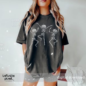 May include: Dark gray t-shirt featuring a white graphic of three dancing skeletons with pumpkin heads. The shirt has short sleeves and a relaxed fit. The text "digital download" is visible on the bottom right.