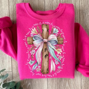 May include: A pink sweatshirt with a graphic of a wooden cross with a pastel pink and blue bow and floral accents. The text "He is risen he is risen he is risen he is risen he is risen he is risen he is risen" surrounds the cross.