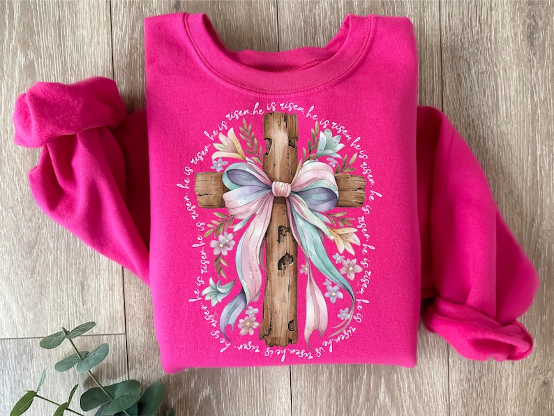 May include: A pink sweatshirt with a graphic of a wooden cross with a pastel pink and blue bow and floral accents. The text "He is risen he is risen he is risen he is risen he is risen he is risen he is risen" surrounds the cross.
