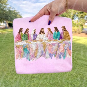May include: A light pink sweatshirt featuring an embroidered depiction of the Last Supper. The artwork showcases figures in colorful robes seated at a table with intricate, flowing details. The sweatshirt is held up by a hand with blue nail polish.