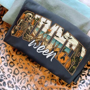 May include: Dark gray t-shirt with the words "Summerween" in a stylized design. The design incorporates a skeleton, beach scene, bats, and pumpkins, blending summer and Halloween themes.