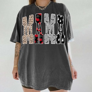 May include: Dark gray t-shirt with the word "MAMA" in a bold, decorative font. The letters feature various patterns, including leopard print, cherries, checkered, bats, and ghosts. The shirt has short sleeves and a relaxed fit.