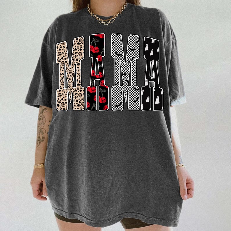 May include: Dark gray t-shirt with the word "MAMA" in a bold, decorative font. The letters feature various patterns, including leopard print, cherries, checkered, bats, and ghosts. The shirt has short sleeves and a relaxed fit.