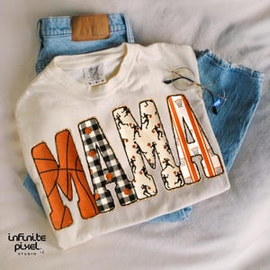 Puede incluir: Sudadera de color crema con la palabra "MAMA" en letras grandes y decorativas. La "M" es naranja con un dise&ntilde;o de baloncesto, la "A" es naranja con un dise&ntilde;o de canasta de baloncesto, y las otras letras tienen patrones de baloncesto y jugadores. La sudadera se combina con vaqueros azules y gafas.