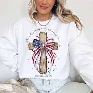 K&ouml;nnte beinhalten: Wei&szlig;es Sweatshirt mit einem Design aus einem verwitterten Holzkreuz. Das Kreuz ist mit einer rot-wei&szlig;-blauen Schleife und einer amerikanischen Flagge verziert. Der Text "Wo der Geist des Herrn ist, da ist Freiheit" ist um das Kreuz herum gedruckt. Der Text "Korinther 3:17" ist unter dem Kreuz gedruckt.