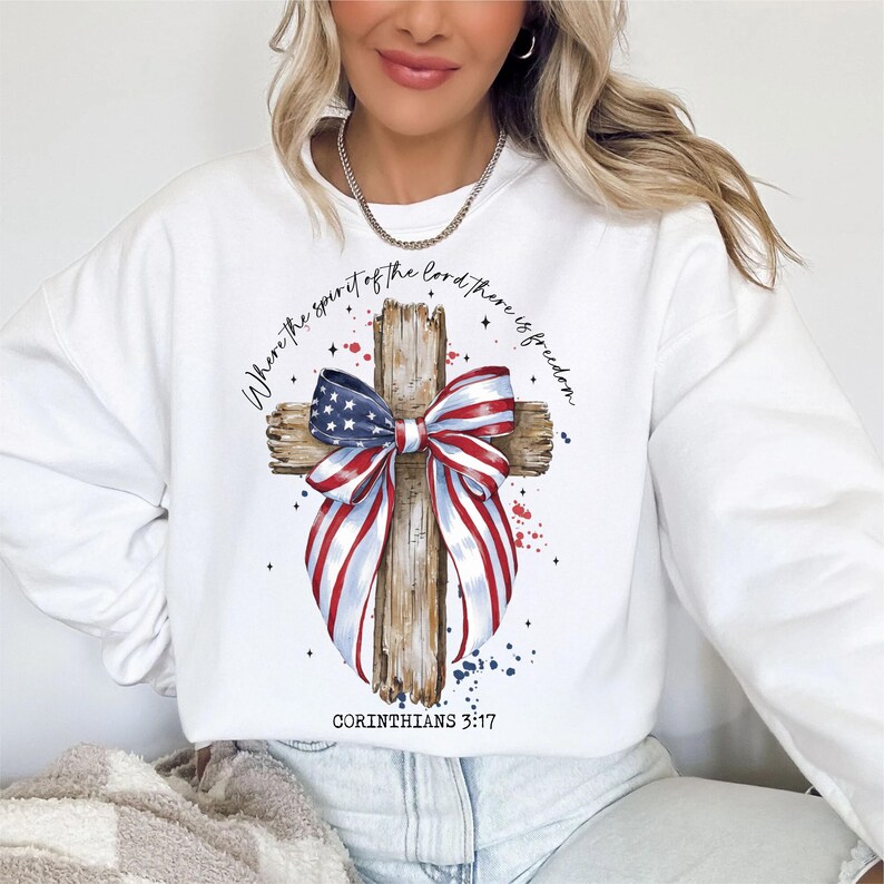 K&ouml;nnte beinhalten: Wei&szlig;es Sweatshirt mit einem Design aus einem verwitterten Holzkreuz. Das Kreuz ist mit einer rot-wei&szlig;-blauen Schleife und einer amerikanischen Flagge verziert. Der Text "Wo der Geist des Herrn ist, da ist Freiheit" ist um das Kreuz herum gedruckt. Der Text "Korinther 3:17" ist unter dem Kreuz gedruckt.