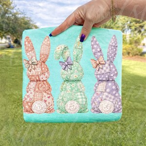 May include: A decorative, square pillow featuring three patchwork bunny rabbits in shades of pink, green, and purple. Each bunny has a bow and a fluffy white tail, set against a turquoise background.
