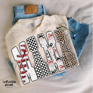May include: A cream-coloured t-shirt displaying the word "MAMA" in a bold, distressed font. The letters feature diverse patterns: red glitter stripes, black and white checkered, baseballs with red bows, and leopard print. The shirt is placed with blue jeans.