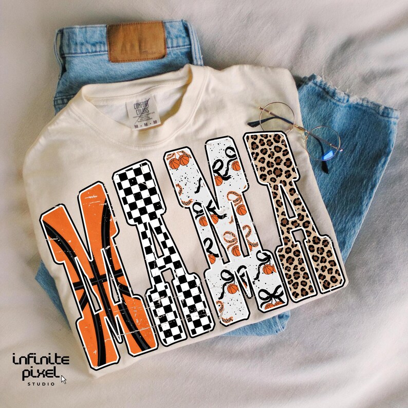 Puede incluir: Camiseta color crema con la palabra "MAMA" en un dise&ntilde;o tem&aacute;tico de baloncesto. Las letras presentan balones de baloncesto, un patr&oacute;n de cuadros y un estampado de leopardo. La camiseta est&aacute; sobre unos vaqueros azules.