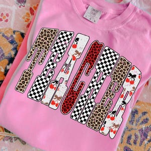 May include: Pink Comfort Colors sweatshirt with the word "PEACHED" in a bold, playful design. The letters feature a mix of leopard print, checkered patterns, and cherry accents, creating a fun and eye-catching aesthetic.