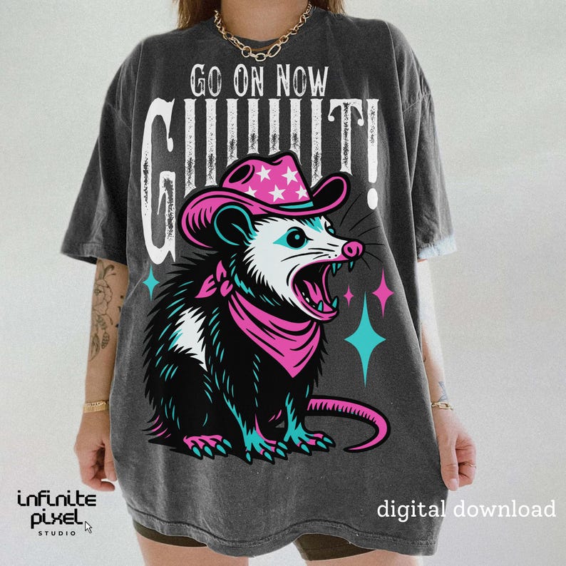 May include: Dark gray t-shirt featuring a cartoon opossum wearing a pink cowboy hat and bandana, with the text "GO ON NOW GUILTY!". The opossum has an open mouth and is surrounded by pink and teal stars.