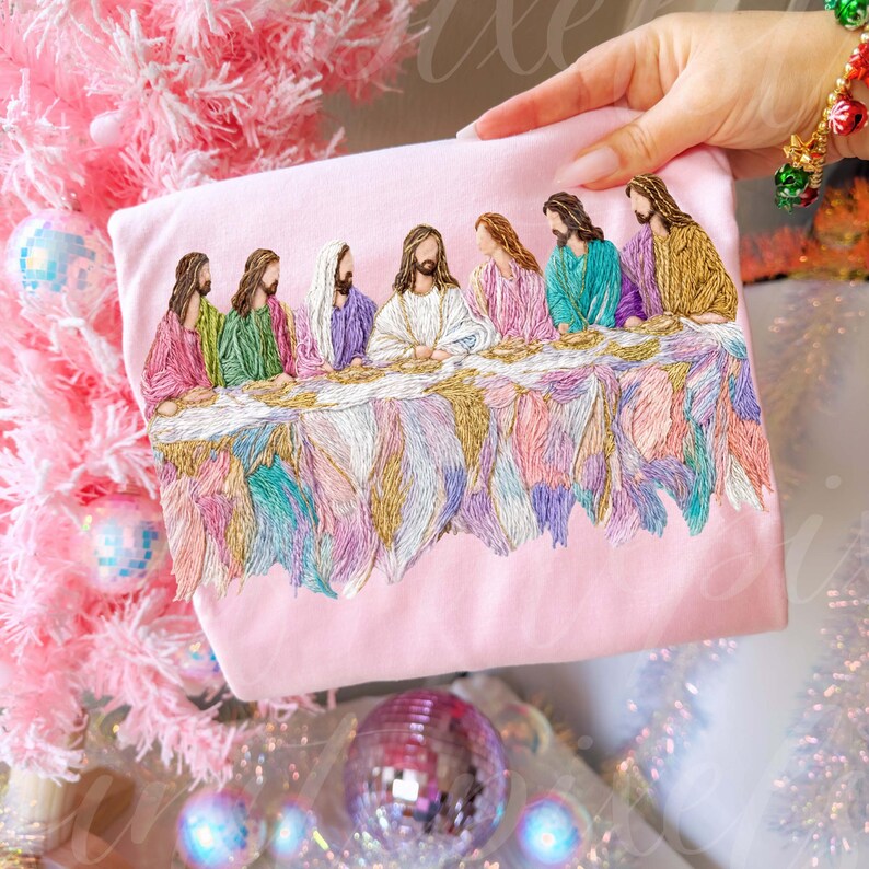 May include: A light pink t-shirt featuring an embroidered depiction of the Last Supper. The embroidery showcases figures in colorful robes seated at a table with a detailed, textured design. The background includes a pink Christmas tree and ornaments.