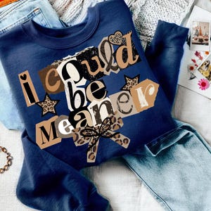 May include: A navy blue sweatshirt with the text "I could be meaner" in a collage style. The design includes leopard print stars and a bow. The sweatshirt is laid on top of a pair of light blue jeans.