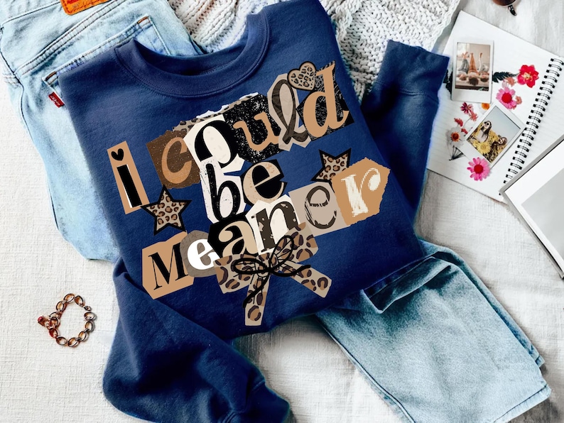 May include: A navy blue sweatshirt with the text "I could be meaner" in a collage style. The design includes leopard print stars and a bow. The sweatshirt is laid on top of a pair of light blue jeans.
