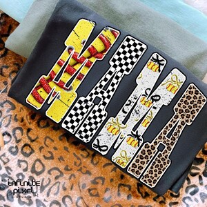 May include: A dark gray t-shirt with the word "WAKE" in a bold, stylized font. Each letter features a unique design: yellow with red snakeskin, black and white checkered, white with gold accents, and leopard print.