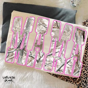 May include: A cream-colored t-shirt with the word "MAMA" in large, pink outlined letters. The letters are filled with a camouflage pattern of white, gray, and brown branches and leaves. The shirt is laying on a dark gray surface.