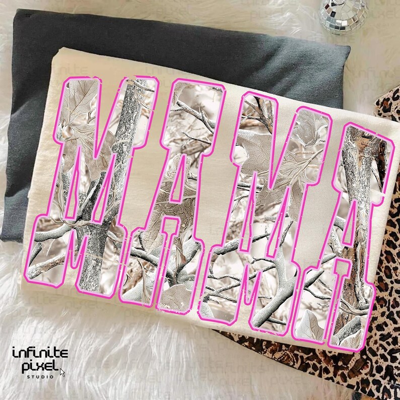 May include: A cream-colored t-shirt with the word "MAMA" in large, pink outlined letters. The letters are filled with a camouflage pattern of white, gray, and brown branches and leaves. The shirt is laying on a dark gray surface.