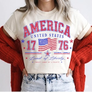 May include: Cream-colored t-shirt featuring a distressed red and blue graphic that reads "AMERICA UNITED STATES 17 76" with a waving American flag. The shirt also includes the text "Land of Liberty" and "Sweet Land of Liberty".