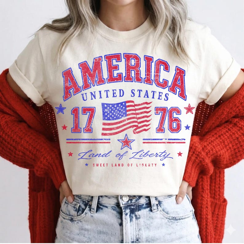 May include: Cream-colored t-shirt featuring a distressed red and blue graphic that reads "AMERICA UNITED STATES 17 76" with a waving American flag. The shirt also includes the text "Land of Liberty" and "Sweet Land of Liberty".