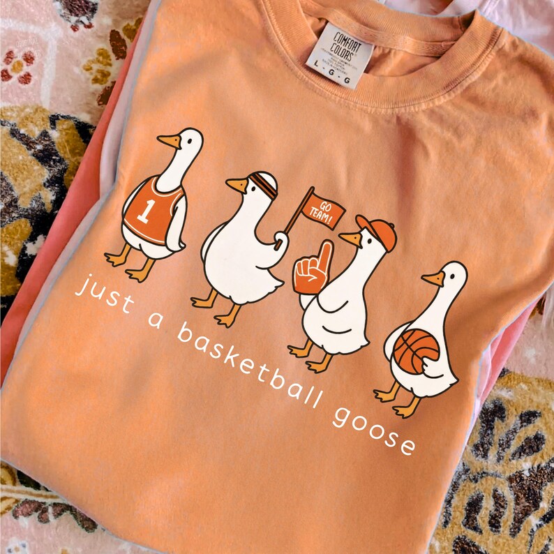 May include: Orange Comfort Colors t-shirt with a cartoon of four geese dressed as basketball fans. The geese are wearing a jersey, headband, hat, and holding a flag, foam finger, and basketball. The text "just a basketball goose" is printed below.