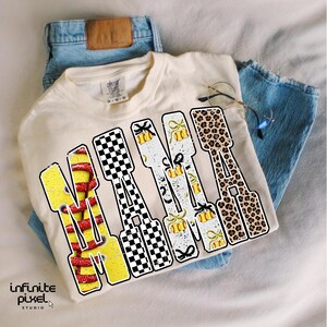 May include: Cream-colored t-shirt with the word "NANA" in a bold, multi-patterned design. The letters feature a mix of yellow, red, black and white patterns, including a checkerboard, leopard print, and softball graphics. The shirt is paired with blue jeans.