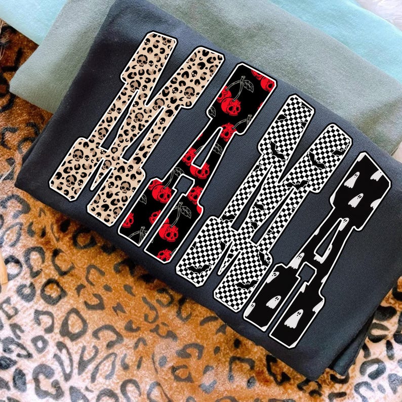 May include: Dark gray sweatshirt with the word "MAMA" in a bold, stylized font. Each letter features a unique pattern: leopard print with skulls, red cherries, black and white checkerboard with bats, and white ghosts.