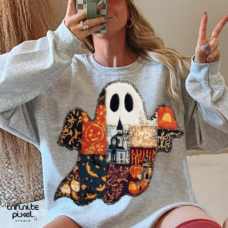 Puede incluir: Una sudadera gris claro con un dise&ntilde;o de fantasma de patchwork. El fantasma est&aacute; hecho de varias telas estampadas en naranja, negro y blanco, con un contorno cosido. El dise&ntilde;o incluye una calabaza y una casa embrujada. El texto "infinite pixel studio" est&aacute; en la parte inferior.