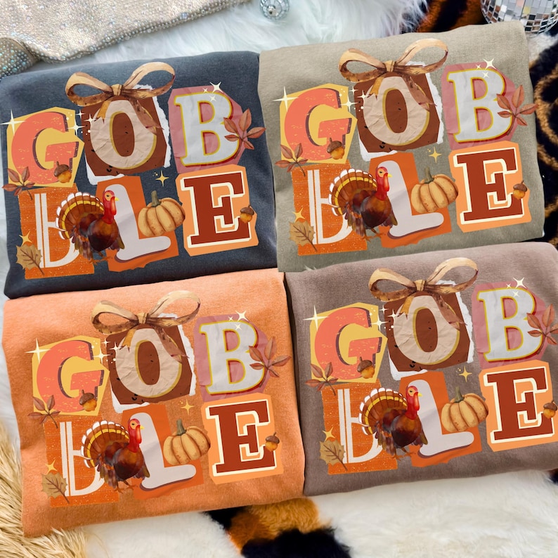 May include: Four crew neck t-shirts in various colors, each featuring the word "GOBBLE" in a fall-themed design. The design includes a turkey, pumpkins, and autumn leaves. The shirts are in shades of gray, tan, orange, and brown.