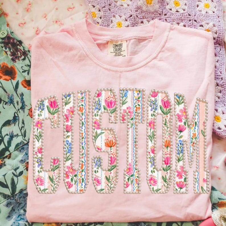 Puede incluir: Una camiseta rosa claro Comfort Colors con la palabra "CUSTOM" en un dise&ntilde;o floral. Las letras est&aacute;n rellenas con un patr&oacute;n floral colorido y delineadas con peque&ntilde;as cuentas perladas. La camiseta est&aacute; doblada sobre una superficie con estampado floral.